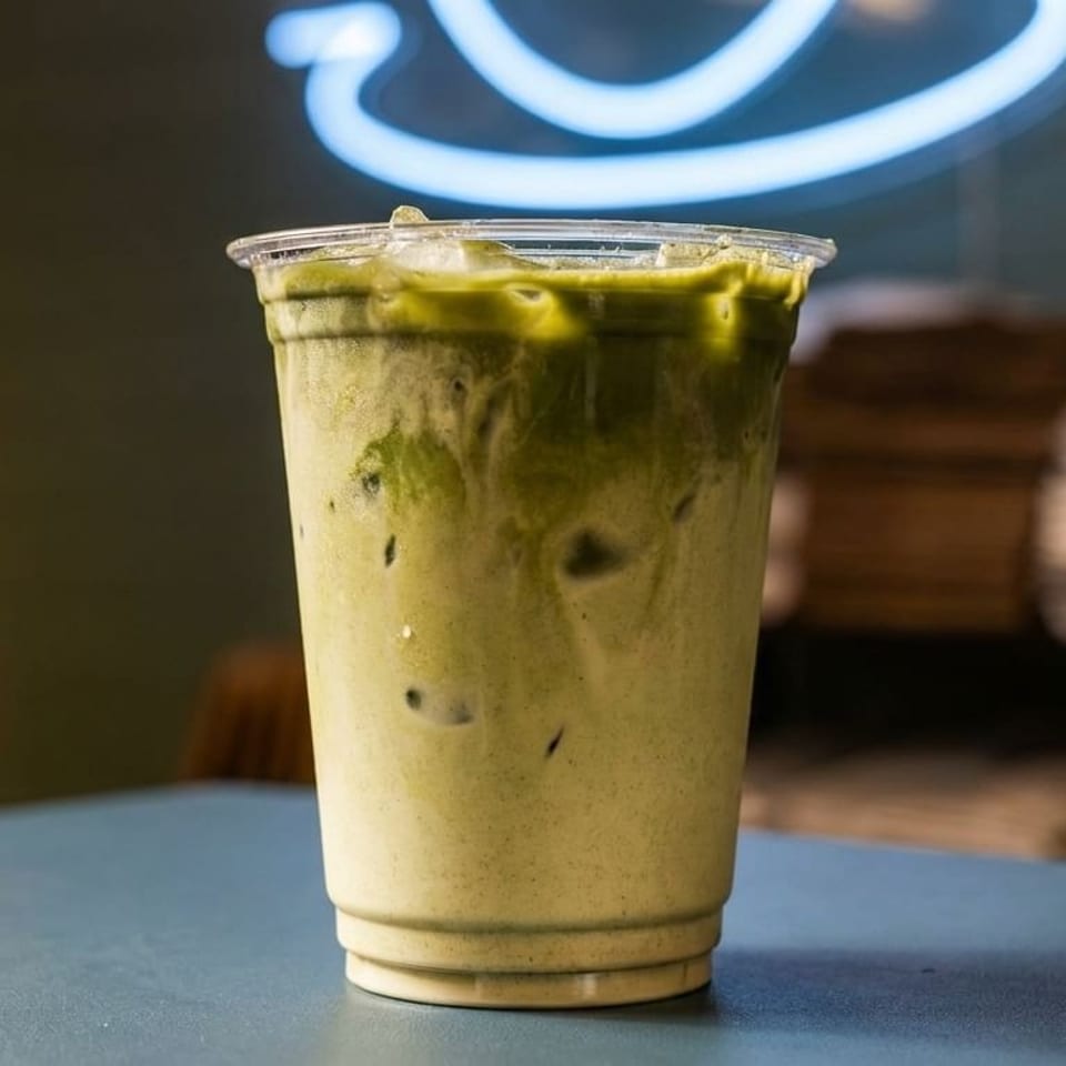 Iced matcha latte in a clear plastic cup on a blue table with a neon sign in the background.